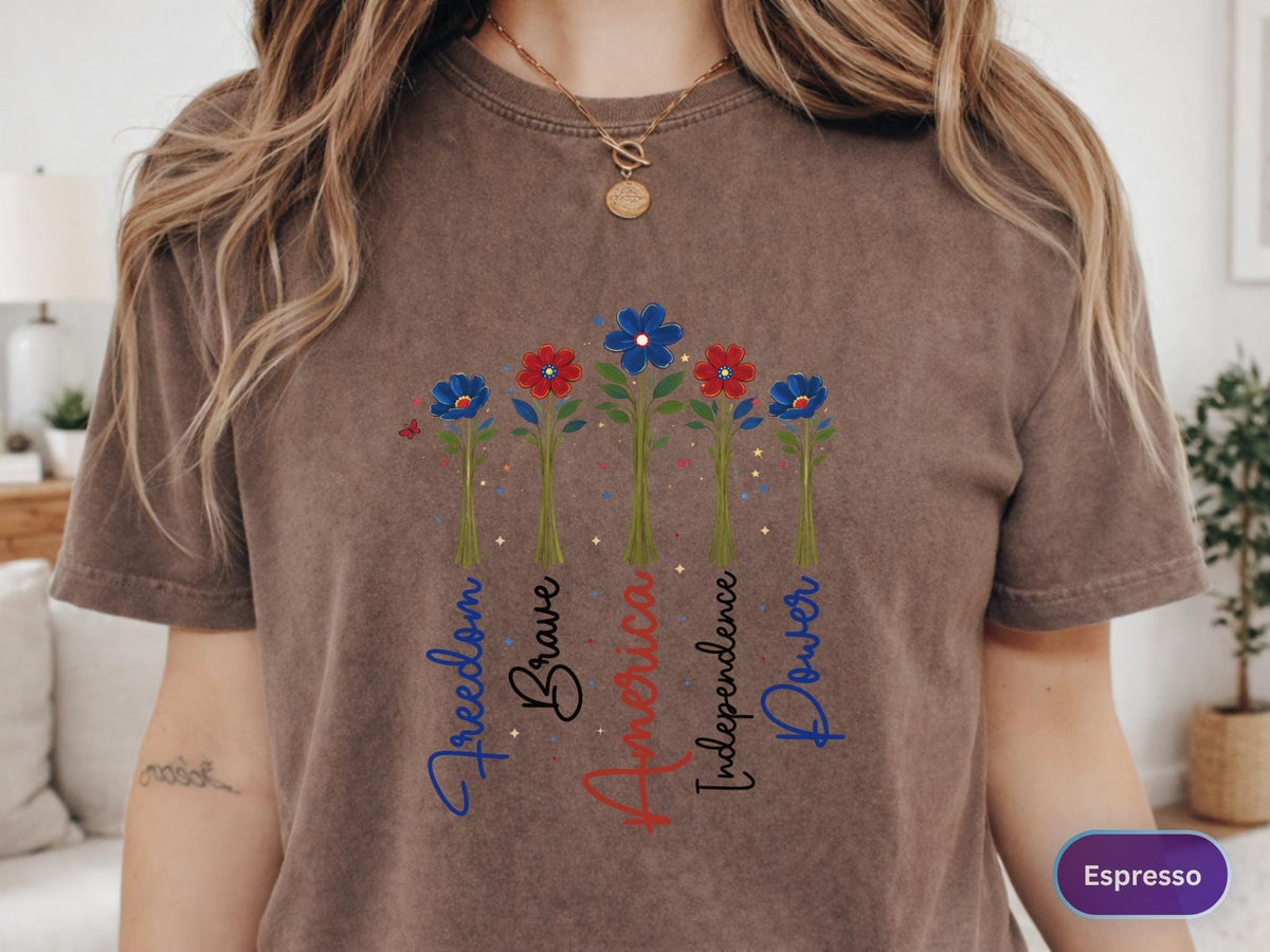 May include: Espresso-colored t-shirt featuring a floral design with red and blue flowers. The words "Freedom," "Brave," "America," "Independence," and "Power" are written vertically below the flowers. The shirt has short sleeves.