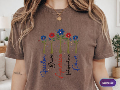 May include: Espresso-colored t-shirt featuring a floral design with red and blue flowers. The words "Freedom," "Brave," "America," "Independence," and "Power" are written vertically below the flowers. The shirt has short sleeves.