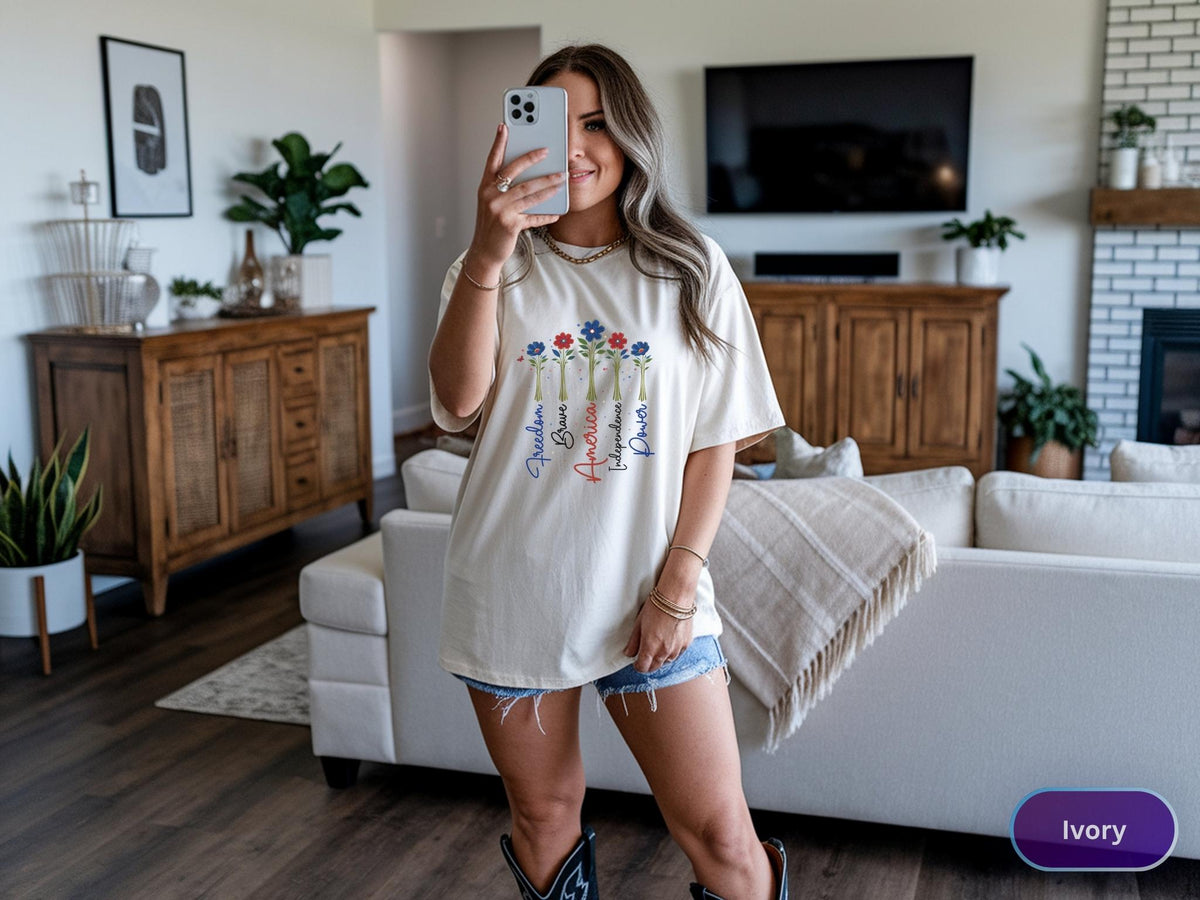May include: Ivory-colored t-shirt with a floral design and the words "Freedom, Brave, America, Independence, Power" in a vertical arrangement. The shirt is worn with denim shorts and cowboy boots.