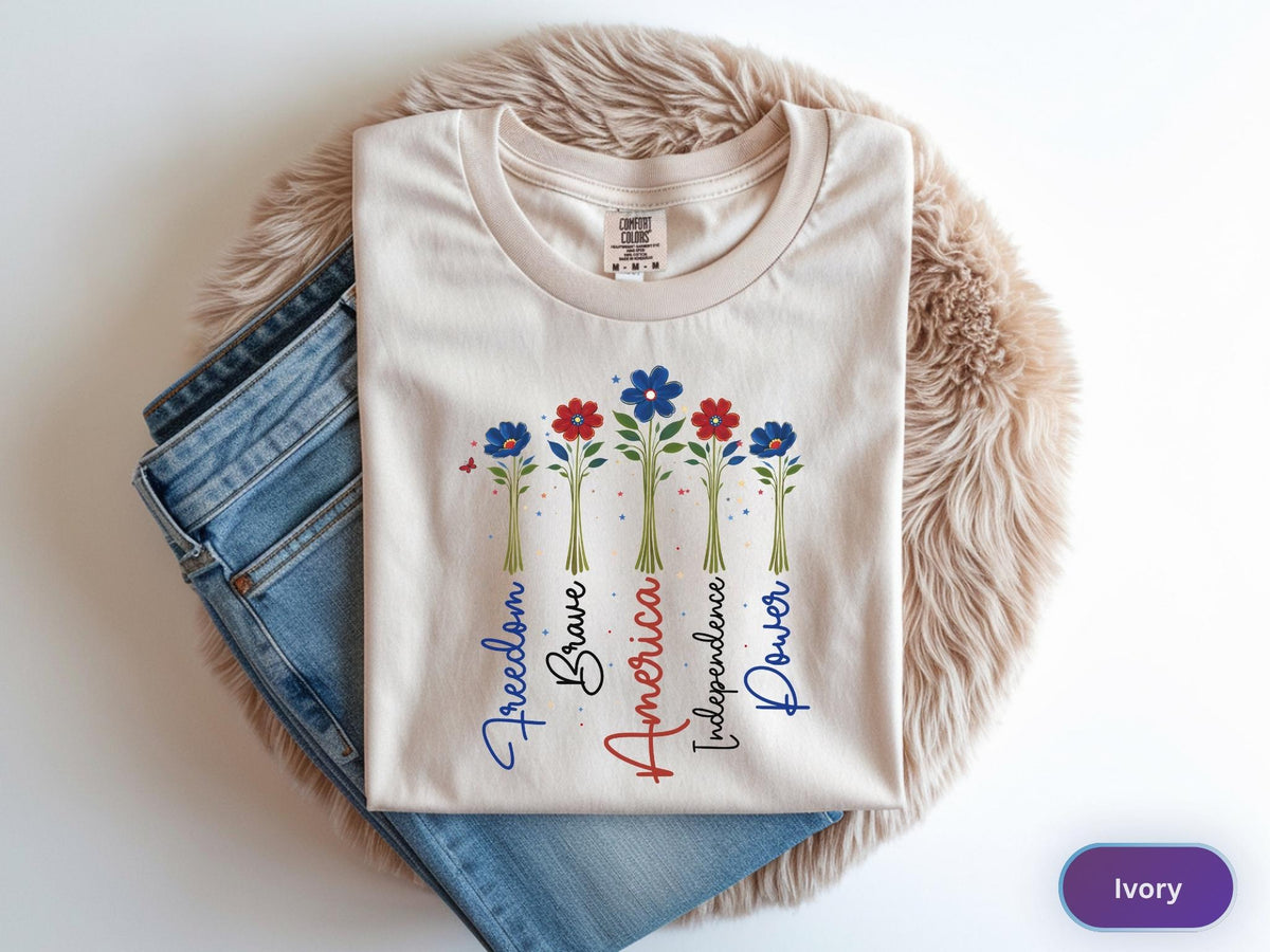 May include: Ivory-colored t-shirt with a floral design in red and blue. The shirt features the words "Freedom, Brave, America, Independence, Power" in a decorative font. The shirt is paired with blue jeans.