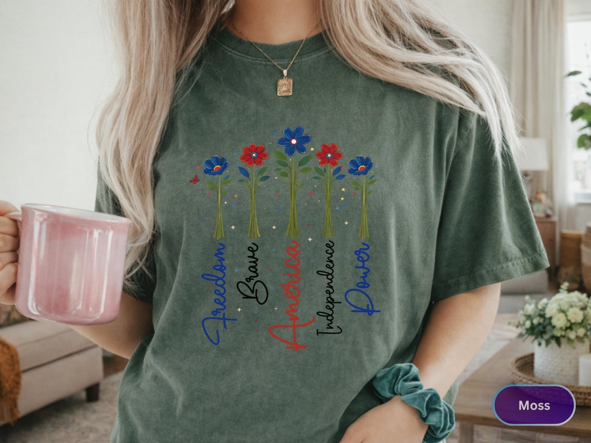 May include: Moss green t-shirt featuring a floral design with red, white, and blue flowers. The words "Freedom, Brave, America, Independence, Power" are written vertically. The shirt is a comfortable, casual style.