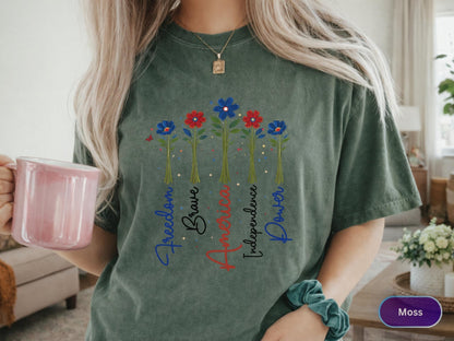May include: Moss green t-shirt featuring a floral design with red, white, and blue flowers. The words "Freedom, Brave, America, Independence, Power" are written vertically. The shirt is a comfortable, casual style.