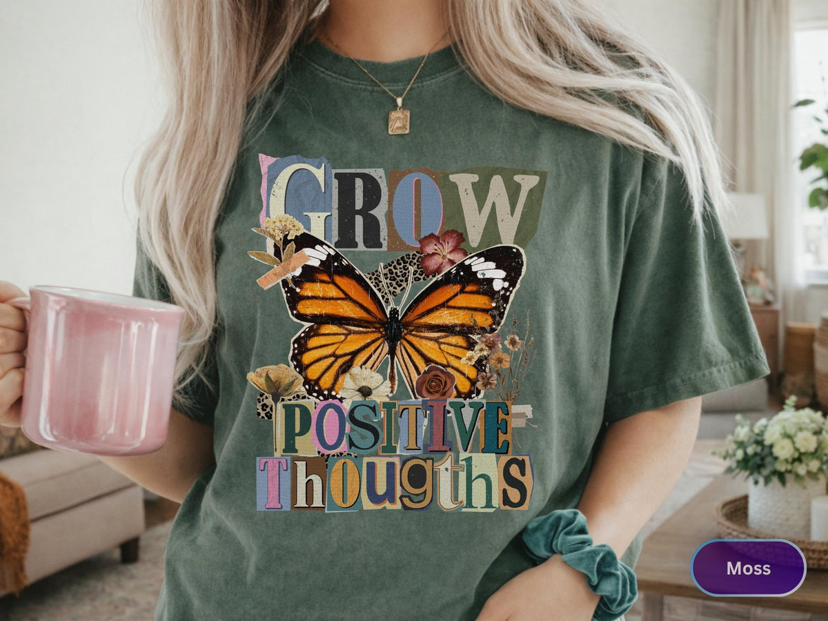 May include: Moss green t-shirt featuring a large butterfly graphic with the words "GROW POSITIVE THOUGHTS" in a colorful, vintage-style font. The butterfly has orange and black wings. A pink mug is held in the hand.