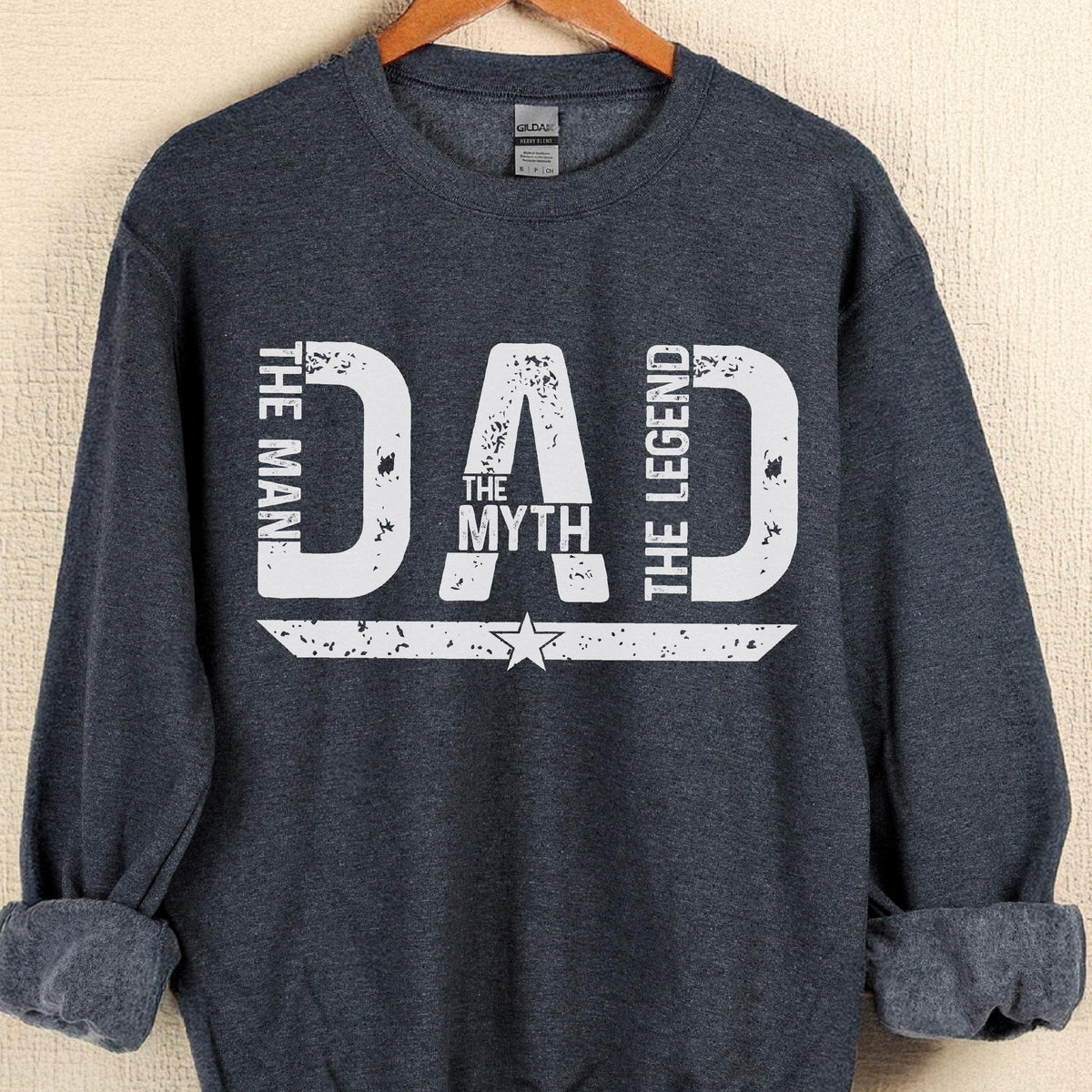 May include: A navy blue sweatshirt with the text "DAD THE MAN THE MYTH THE LEGEND" in distressed white lettering. The design includes a star and a horizontal stripe. The sweatshirt is hanging on a wooden hanger.
