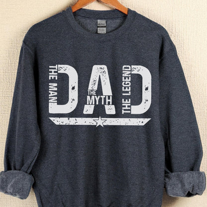 May include: A navy blue sweatshirt with the text "DAD THE MAN THE MYTH THE LEGEND" in distressed white lettering. The design includes a star and a horizontal stripe. The sweatshirt is hanging on a wooden hanger.