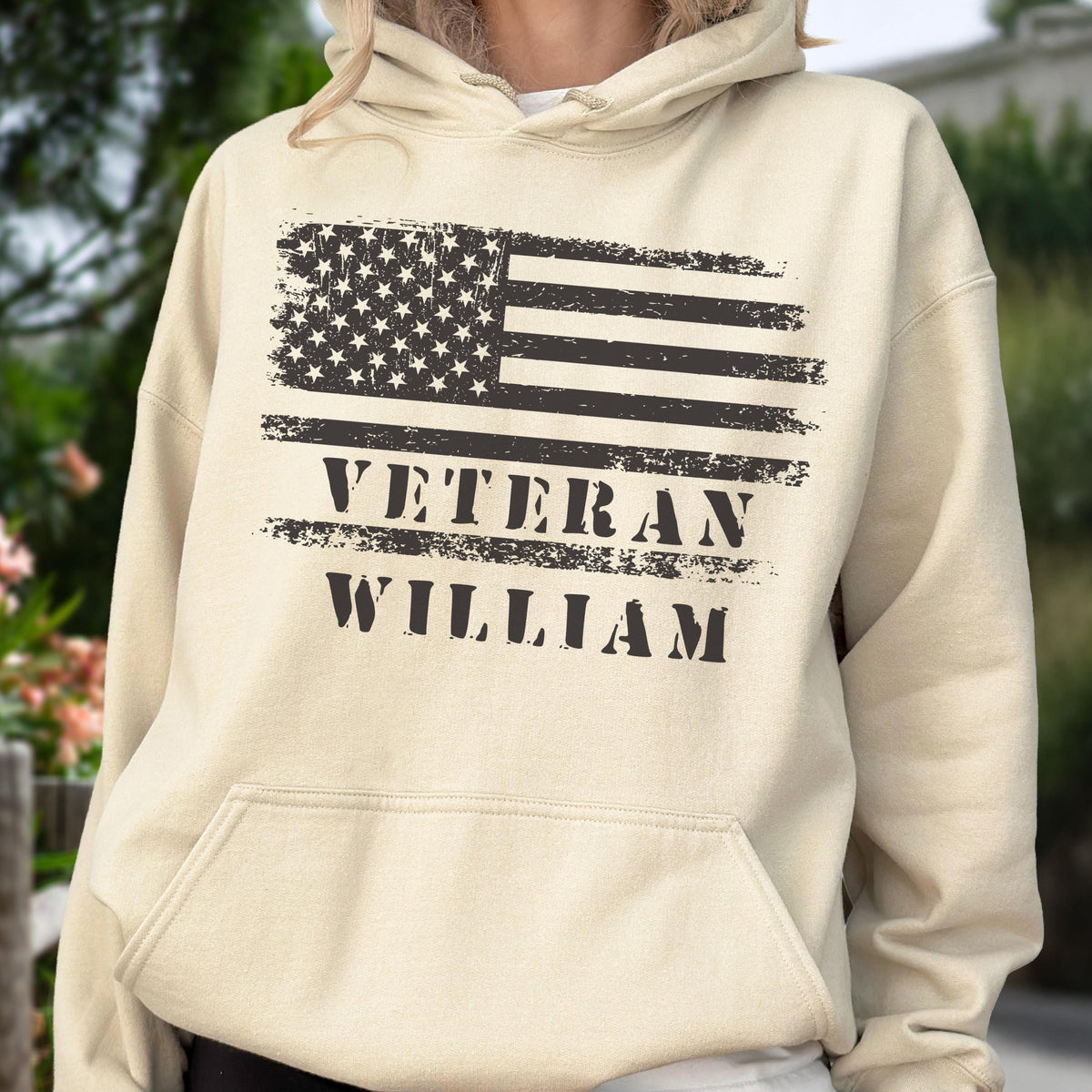 May include: A beige hoodie featuring a distressed American flag design in black. The text "VETERAN WILLIAM" is printed below the flag. The hoodie has a front pocket and a drawstring hood.