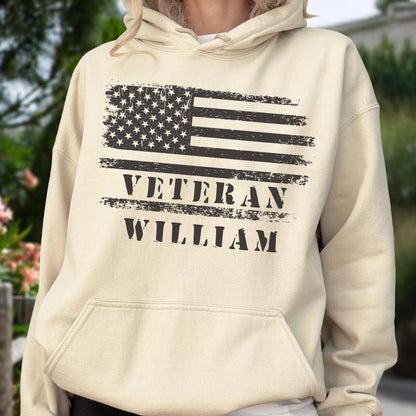 May include: A beige hoodie featuring a distressed American flag design in black. The text "VETERAN WILLIAM" is printed below the flag. The hoodie has a front pocket and a drawstring hood.