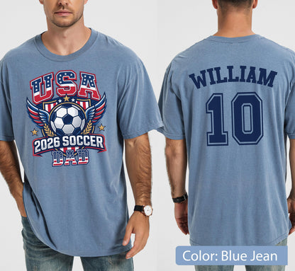 May include: A blue jean-colored t-shirt with a soccer-themed graphic on the front. The graphic includes the text "USA 2026 SOCCER DAD" with a soccer ball and wings. The back of the shirt displays the name "WILLIAM" and the number "10".