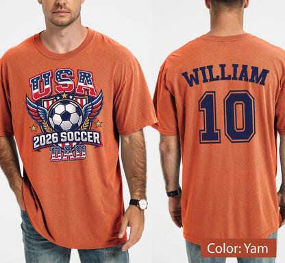 May include: Orange t-shirt with a soccer-themed graphic. The front features "USA" in red and blue, a soccer ball, wings, and "2026 SOCCER DAD." The back displays "WILLIAM" and the number "10" in navy blue.