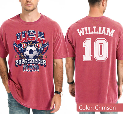 May include: Crimson-colored t-shirt with "USA" and a soccer ball graphic, wings, and stars. The text "2026 SOCCER DAD" is below the ball. The back of the shirt has the name "WILLIAM" and the number "10" in white.