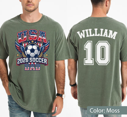 May include: Moss green t-shirt with "USA 2026 SOCCER DAD" graphic on the front, featuring a soccer ball, wings, and stars. The back displays "WILLIAM 10" in white block letters. The shirt is a casual, short-sleeved style.