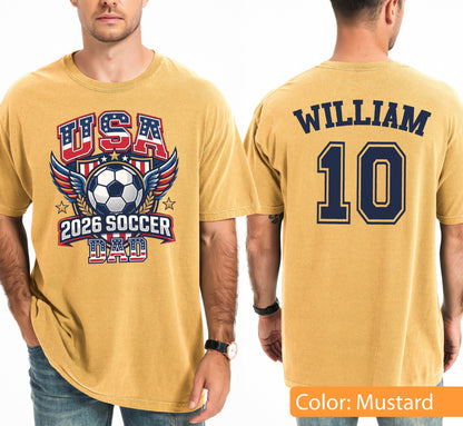 May include: Mustard-colored t-shirt with a soccer-themed graphic. The front features "USA" in red and blue, a soccer ball, wings, and "2026 SOCCER DAD." The back displays "WILLIAM" above the number "10" in navy blue.