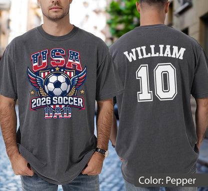 May include: Pepper-colored t-shirt with a soccer-themed graphic on the front, featuring "USA", a soccer ball, and "2026 SOCCER DAD". The back displays "WILLIAM" and the number "10" in white.