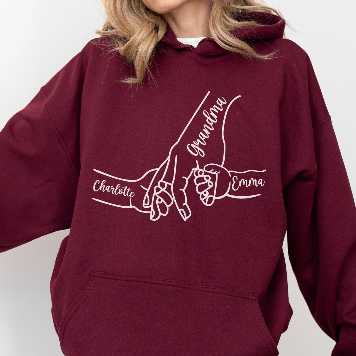 May include: Burgundy hoodie with a white graphic of hands holding each other. The word "Grandma" is written in cursive, with the names "Charlotte" and "Emma" below the hands. The hoodie has a drawstring hood and a front pocket.