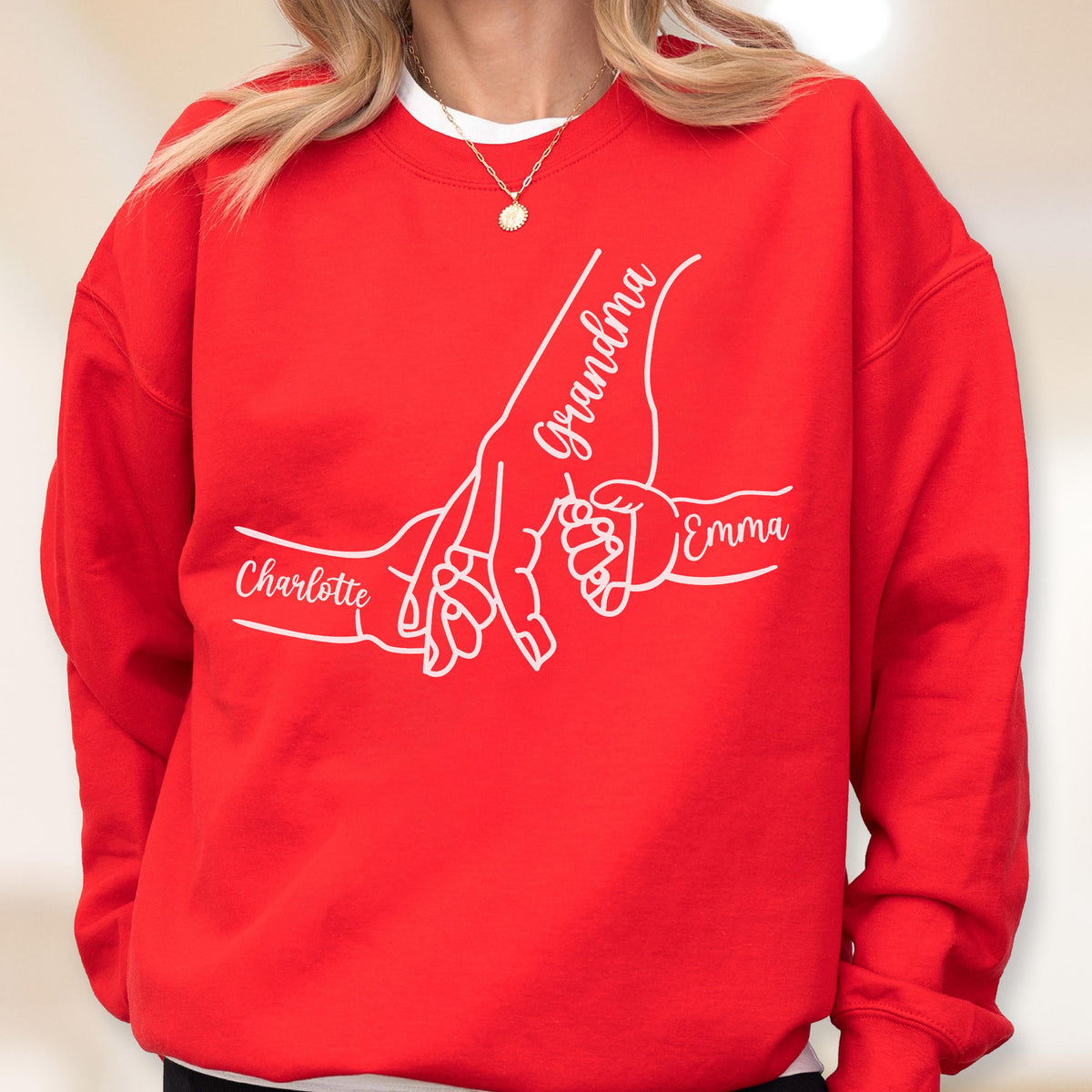 May include: A red sweatshirt with a white graphic of hands holding each other. The word "Grandma" is written on one hand, and the names "Charlotte" and "Emma" are written on the other hands. The sweatshirt is a crew neck style.