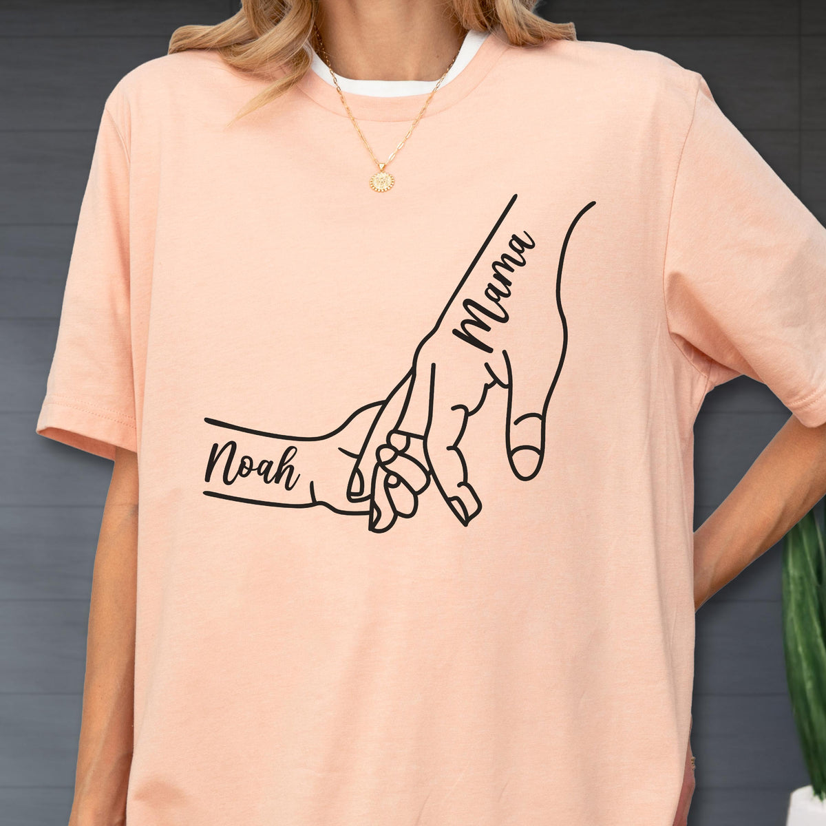 May include: Peach-colored t-shirt with a white collar. The shirt features a black line drawing of two hands holding each other. The word "Mama" is written on one hand, and the name "Noah" is written on the other.