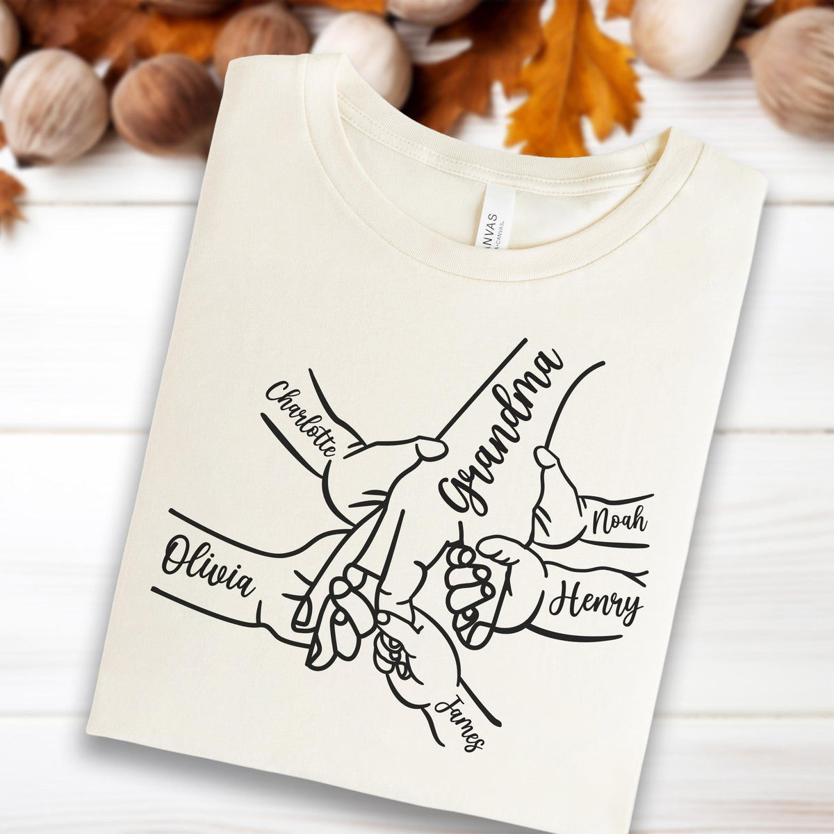 May include: Cream-colored t-shirt featuring a black graphic of hands holding each other, with the word "Grandma" and the names "Olivia", "Charlotte", "Noah", "Henry", and "James" printed on it.