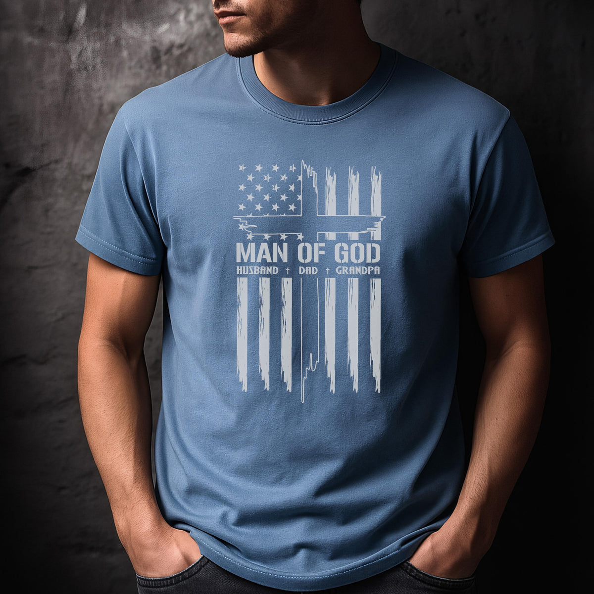 May include: A light blue t-shirt with a white graphic design. The design features an American flag with a cross and the words "MAN OF GOD", "HUSBAND", "DAD", and "GRANDPA".