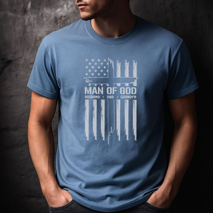 May include: A light blue t-shirt with a white graphic design. The design features an American flag with a cross and the words "MAN OF GOD", "HUSBAND", "DAD", and "GRANDPA".