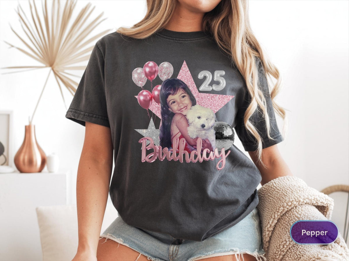 May include: A charcoal gray short-sleeve t-shirt featuring a graphic print of a person holding a white puppy. The design includes a large pink glitter star, silver balloons, the number 25, a disco ball, and the word Birthday in pink script.
