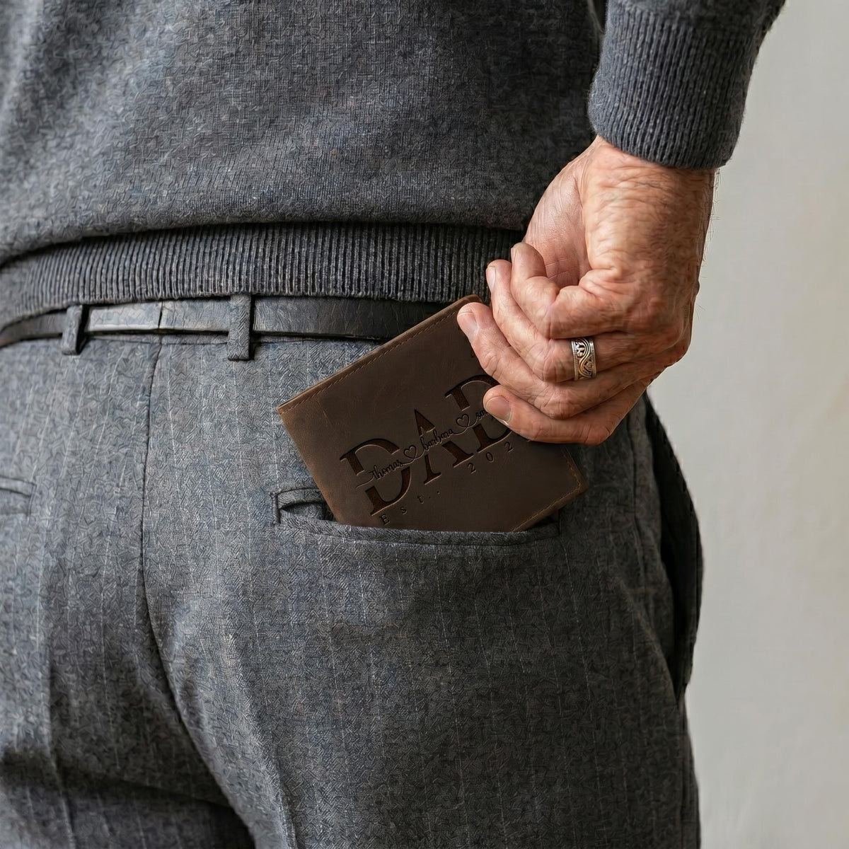 May include: A dark brown leather wallet featuring the embossed text DAD with a small heart symbol between the letters. The wallet is partially tucked into the back pocket of grey pinstriped trousers, with a silver ring visible on a finger.