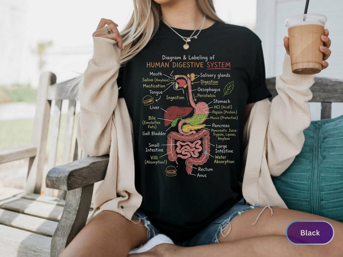 May include: A black t-shirt featuring a colorful anatomical diagram of the human digestive system with labels including mouth, esophagus, stomach, liver, pancreas, and intestines, along with small illustrations of food items and scientific text.