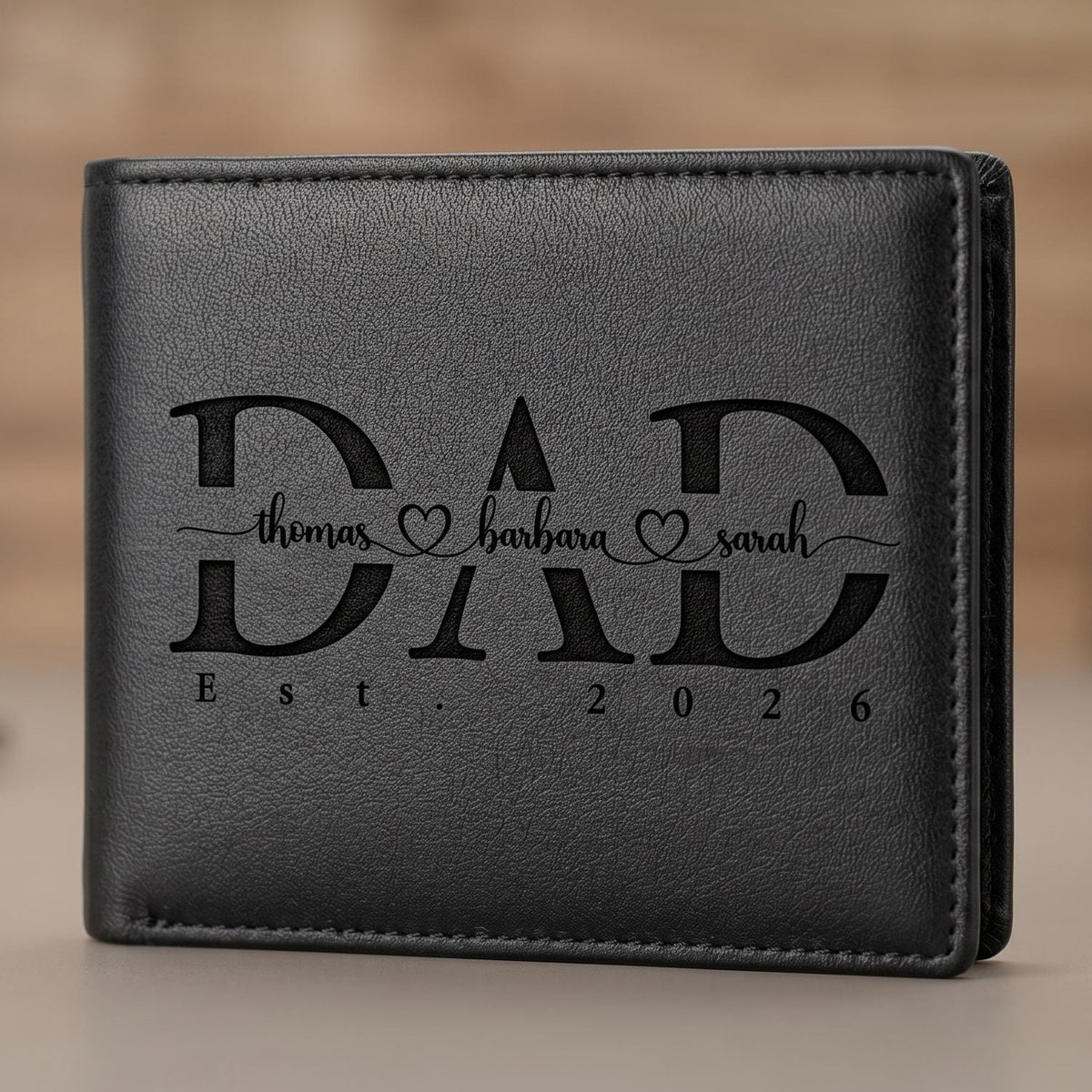 May include: A black bi-fold wallet with a textured surface. The front features the engraved word DAD in large capital letters. Inside the letters, the names thomas, barbara, and sarah are written in script with heart icons. Below is Est. 2026.