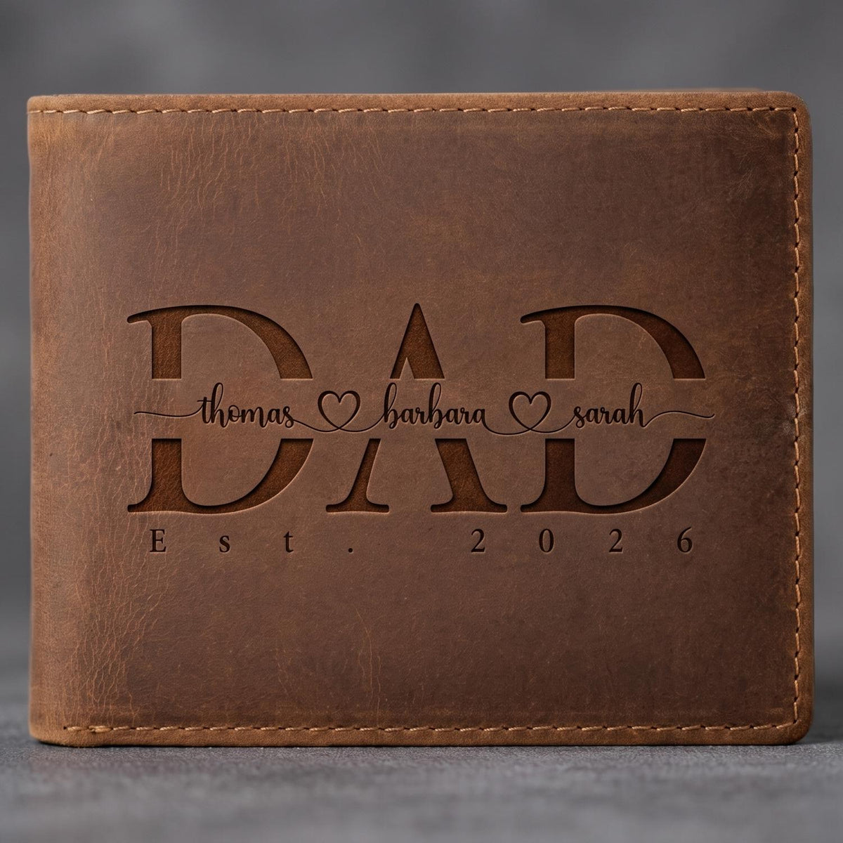 May include: A brown leather bifold wallet featuring the engraved word DAD in large capital letters. The names thomas, barbara, and sarah are written in cursive script between the letters, separated by heart icons. The text Est. 2026 is below.