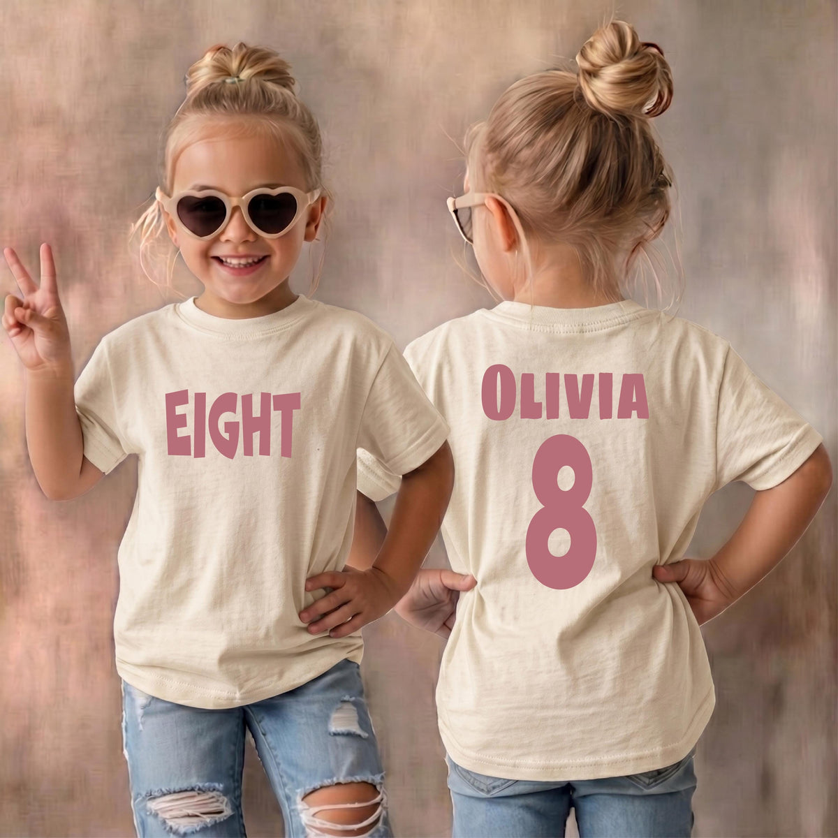 May include: A cream-colored short-sleeve t-shirt featuring the word EIGHT in pink letters on the front and the name OLIVIA with the number 8 in pink on the back. The shirt is worn with blue jeans and heart-shaped sunglasses.