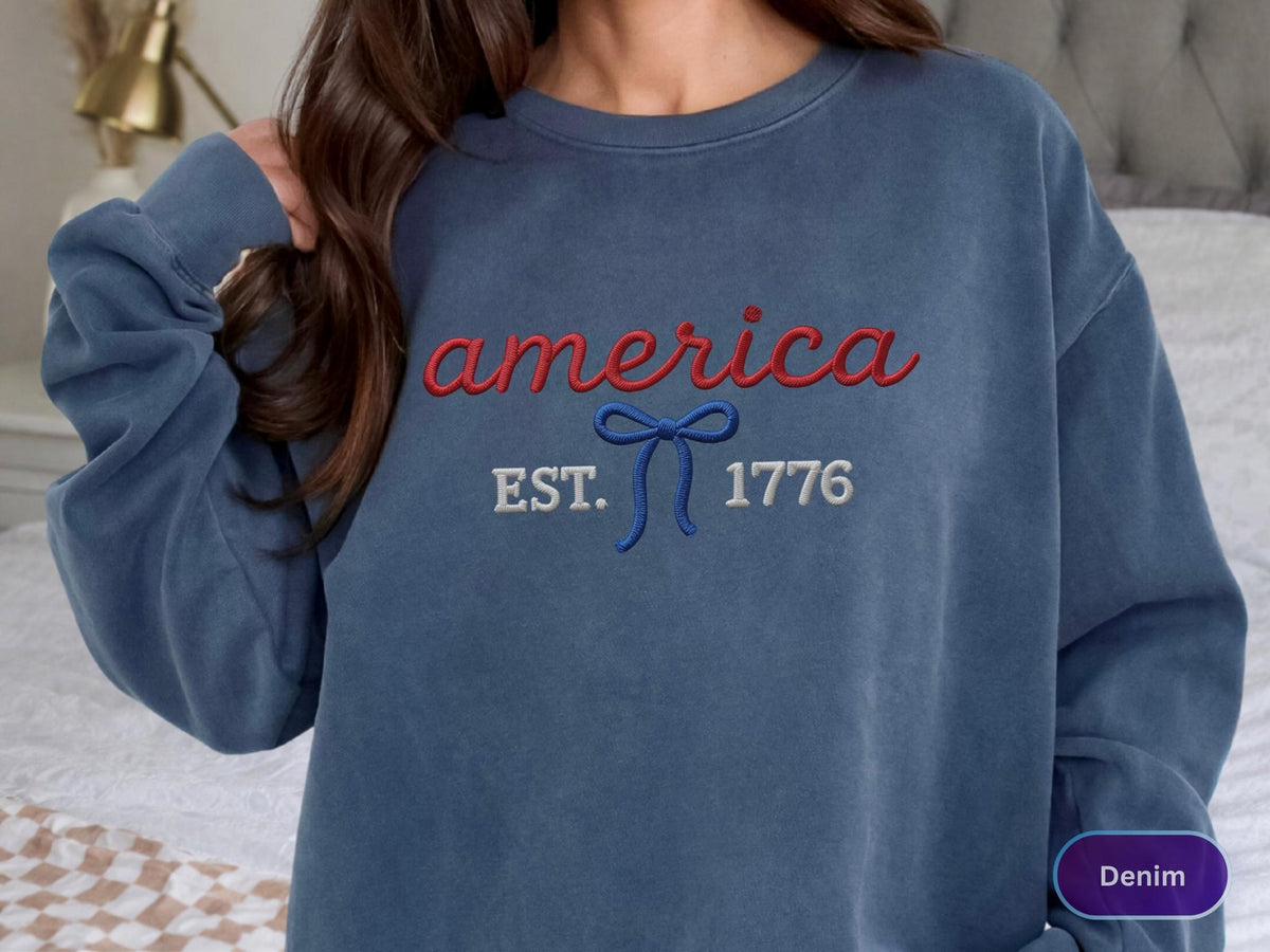 May include: A blue crewneck sweatshirt featuring embroidered text that reads america in red script, with a blue bow icon below it, followed by the white embroidered text EST. 1776. The garment has a long-sleeved design and a solid blue color.