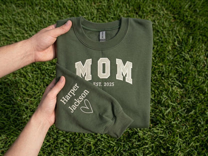 May include: An olive green crewneck sweatshirt featuring the white embroidered text MOM EST. 2025 on the chest. The sleeve is embroidered with the names Harper and Jackson in white, accompanied by a small white heart outline graphic.