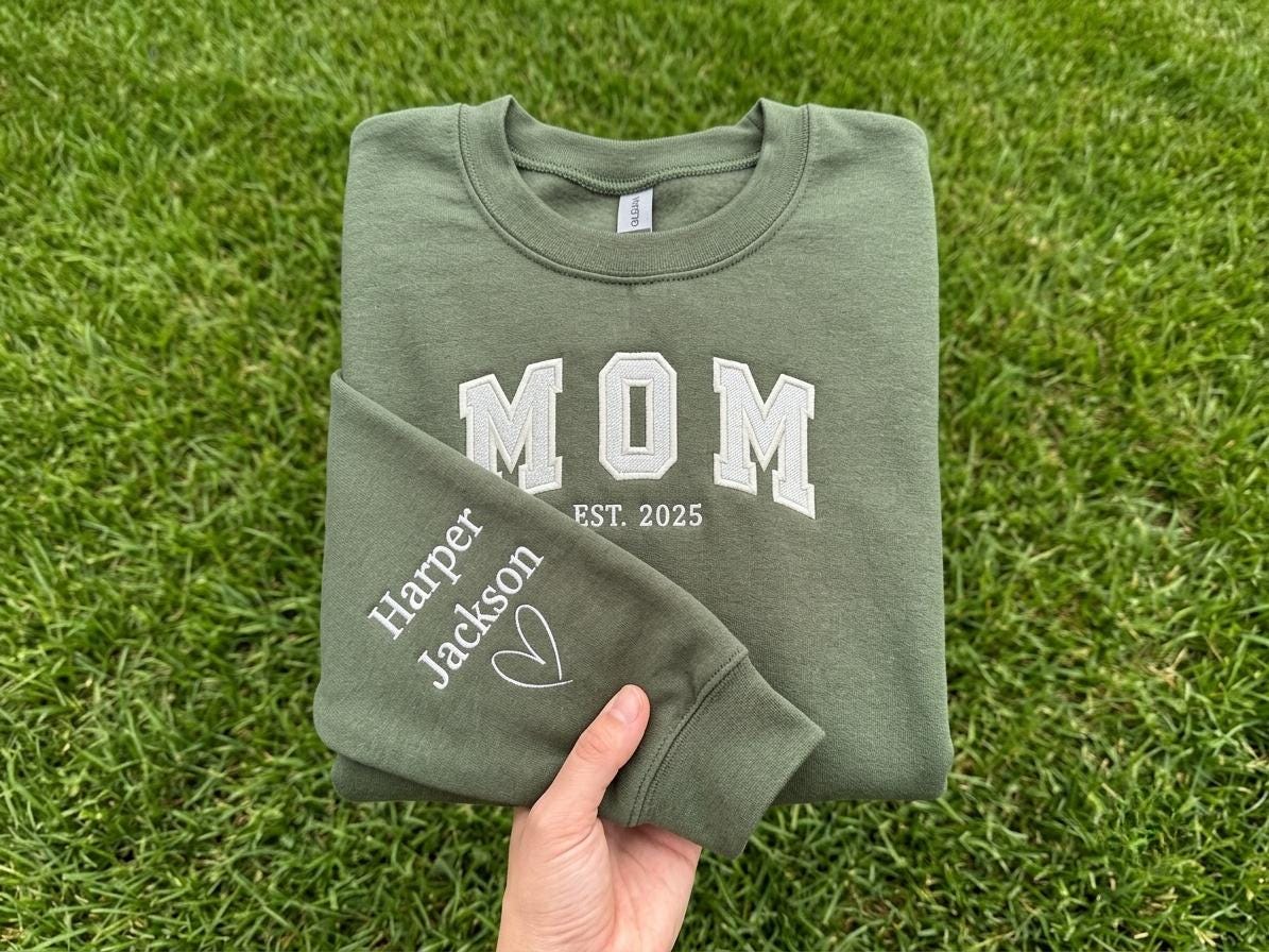 May include: An olive green crewneck sweatshirt featuring white embroidered text that reads MOM EST. 2025 on the chest. The sleeve is embroidered with the names Harper and Jackson followed by a small white heart outline design.