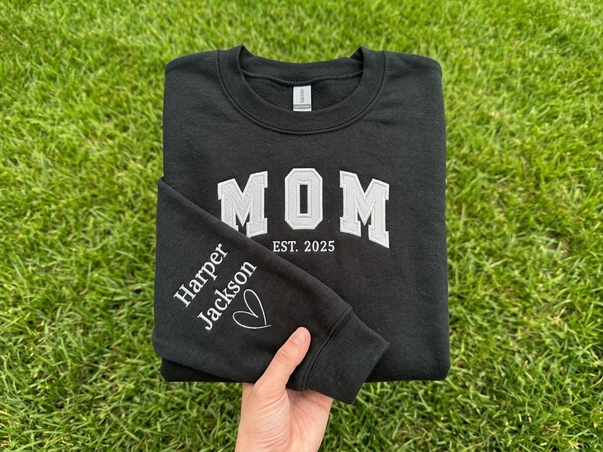 May include: A black crewneck sweatshirt featuring white embroidered text that reads MOM EST. 2025 on the chest. The sleeve is embroidered with the names Harper and Jackson in white, accompanied by a small white heart outline graphic.