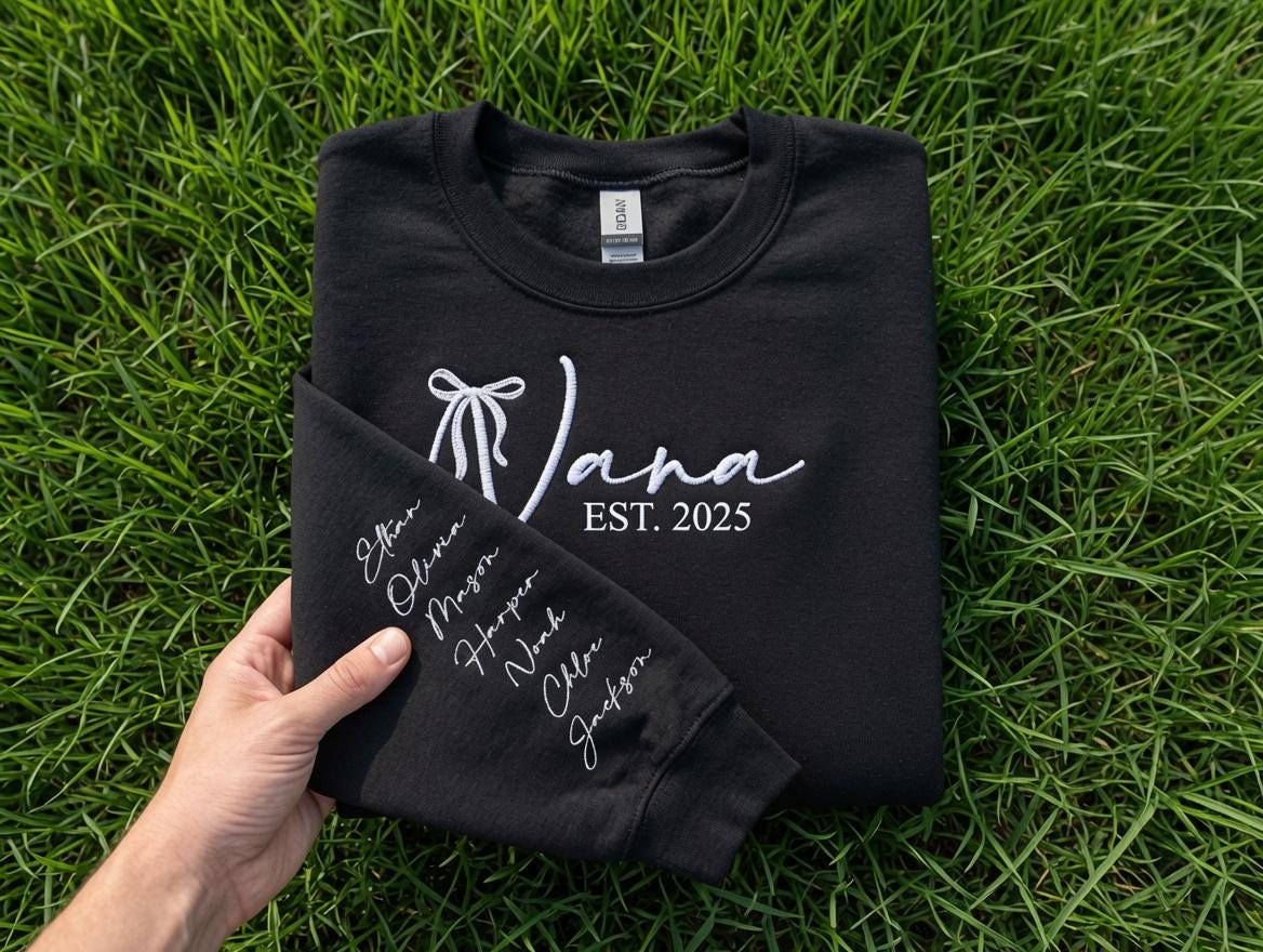 May include: A black crewneck sweatshirt featuring white embroidery on the chest with a bow icon, the name Jana, and the text EST. 2025. The sleeve is folded over to reveal a vertical list of names embroidered in white script: Ethan, Olivia, Mason, Harper, Noah, Chloe, and Jackson.