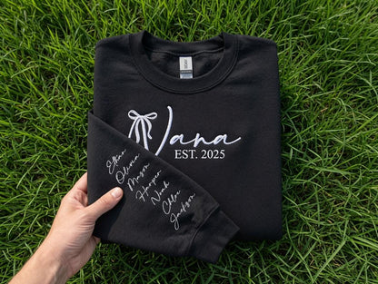 May include: A black crewneck sweatshirt featuring white embroidery on the chest with a bow icon, the name Jana, and the text EST. 2025. The sleeve is folded over to reveal a vertical list of names embroidered in white script: Ethan, Olivia, Mason, Harper, Noah, Chloe, and Jackson.