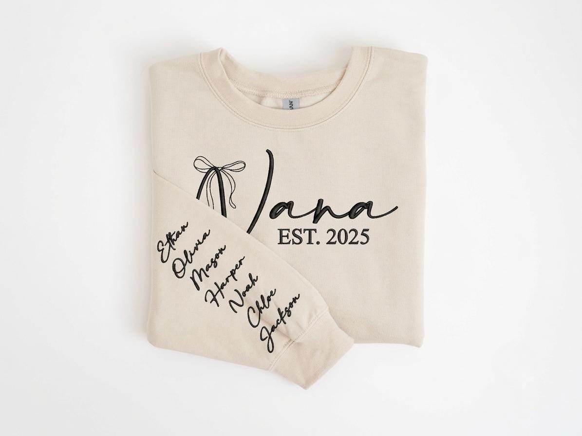 May include: A beige crewneck sweatshirt featuring black embroidered text that reads Nana EST. 2025 with a bow graphic. The sleeve is embroidered with a vertical list of names including Ethan, Olivia, Mason, Harper, Noah, Chloe, and Jackson in black.
