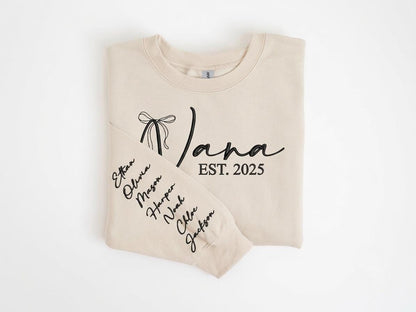 May include: A beige crewneck sweatshirt featuring black embroidered text that reads Nana EST. 2025 with a bow graphic. The sleeve is embroidered with a vertical list of names including Ethan, Olivia, Mason, Harper, Noah, Chloe, and Jackson in black.