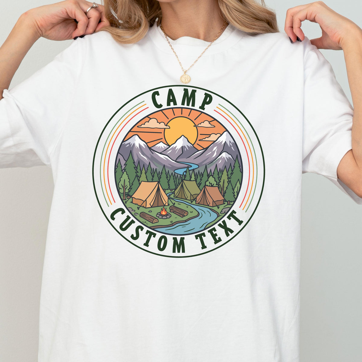 May include: A white t-shirt featuring a circular graphic design of a mountain landscape with a sun, pine trees, a river, and two tents. The text "CAMP" is arched at the top and "CUSTOM TEXT" is arched at the bottom in dark green lettering.