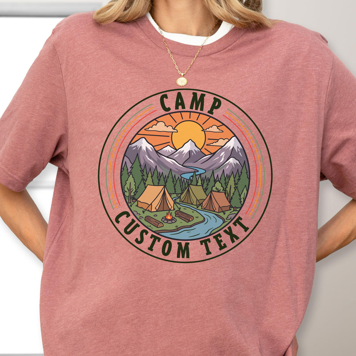 May include: A dusty rose short-sleeve t-shirt featuring a circular graphic print. The design depicts a mountain landscape with a sun, pine trees, tents, a campfire, and a river. The text "CAMP" and "CUSTOM TEXT" is printed in dark green.