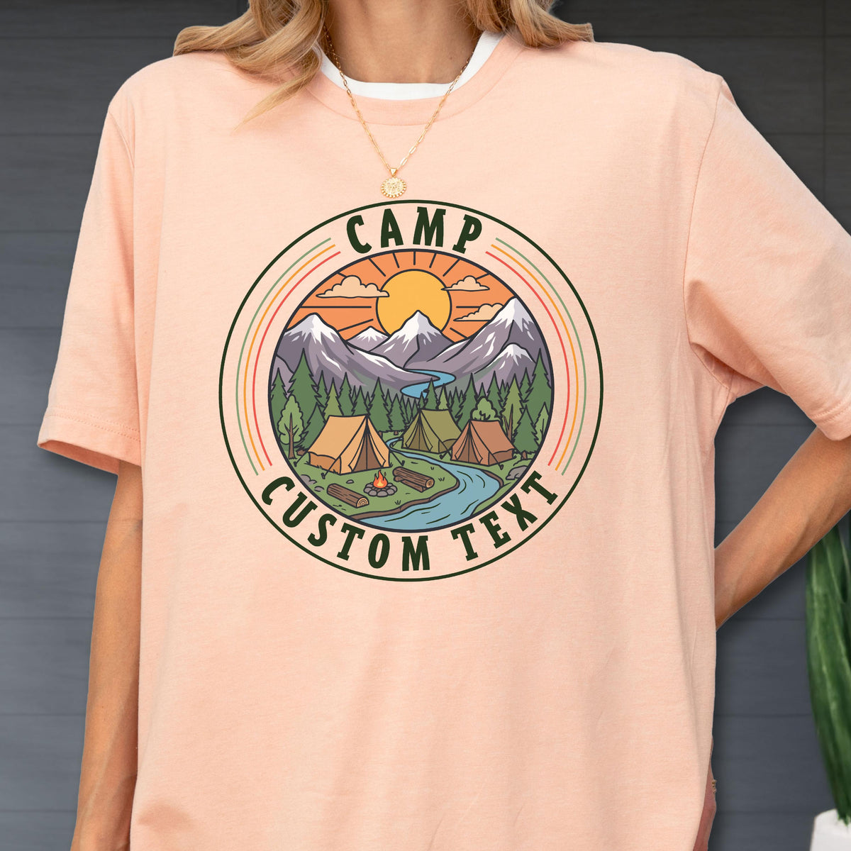 May include: A peach-colored t-shirt featuring a circular graphic with a mountain landscape, pine trees, a river, tents, and a campfire. The text "CAMP" is arched at the top and "CUSTOM TEXT" is arched at the bottom in dark green lettering.