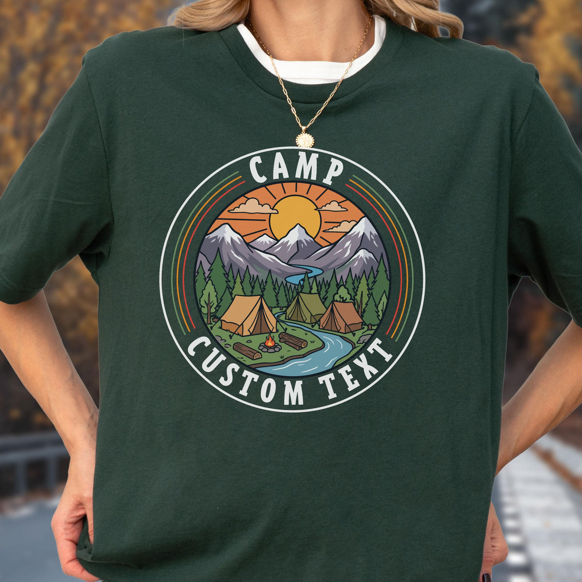 May include: A dark green t-shirt featuring a circular graphic with a mountain landscape, pine trees, a river, and tents. The design includes a yellow sun, a campfire, and the text "CAMP" at the top and "CUSTOM TEXT" at the bottom in white.