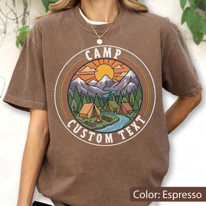 May include: An espresso-colored t-shirt featuring a circular graphic print. The design depicts a mountain landscape with a sun, pine trees, tents, a campfire, and a river. The text CAMP and CUSTOM TEXT are printed in white within the circular border.