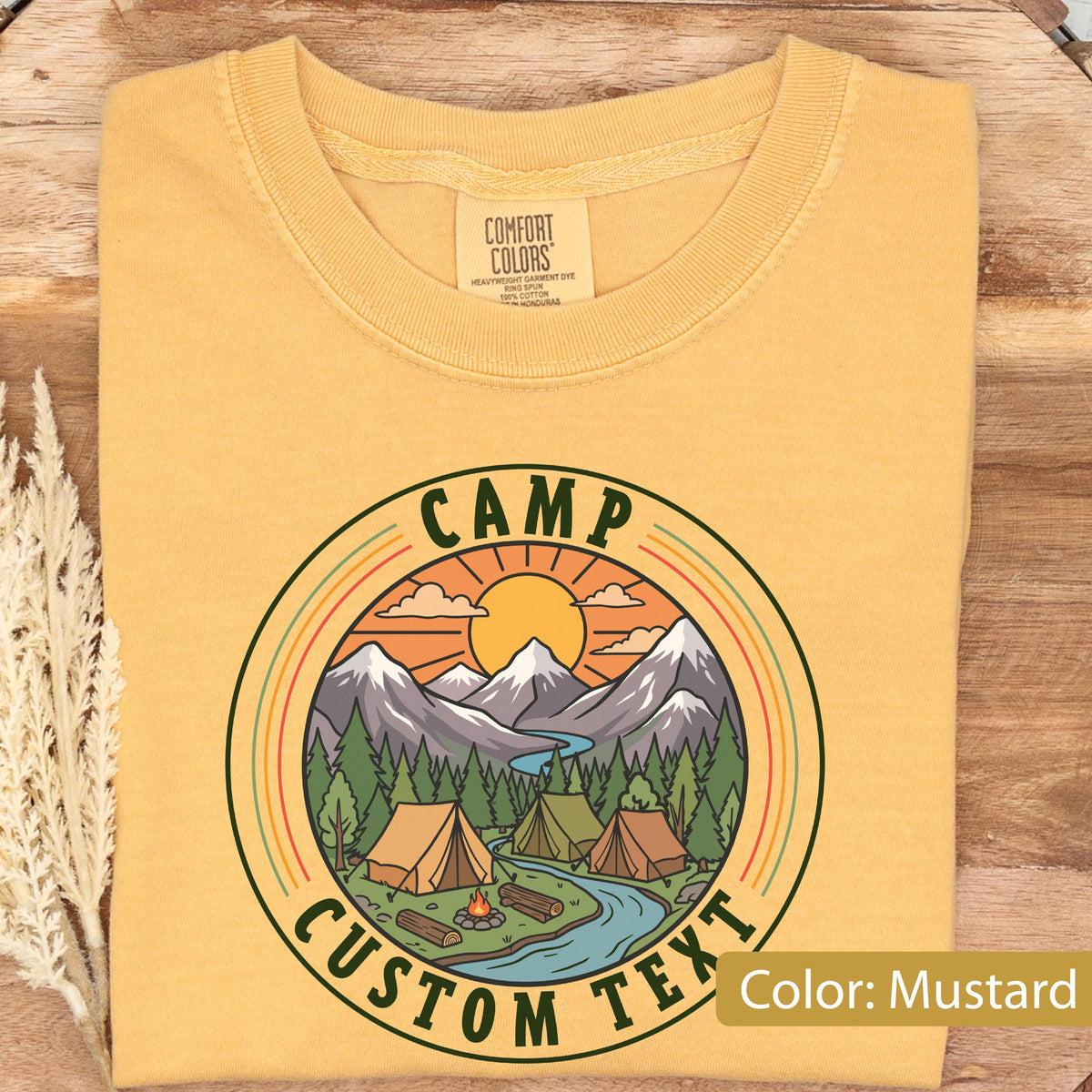 May include: A mustard yellow t-shirt featuring a circular graphic with the text CAMP CUSTOM TEXT. The design depicts a mountain range, a sun, a forest, a river, and two tents. The shirt has a crew neckline and a Comfort Colors brand label.