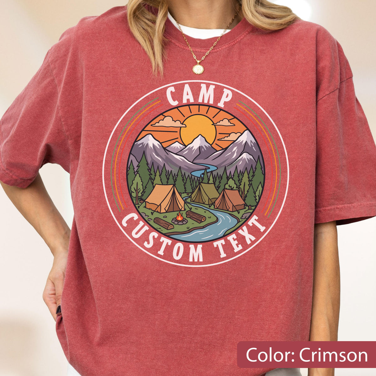 May include: A crimson short-sleeve t-shirt featuring a circular graphic of a mountain landscape with tents, a river, and a campfire. The text CAMP CUSTOM TEXT is printed in white within the circular border, set against a sunset sky background.