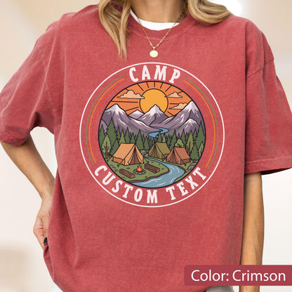 May include: A crimson short-sleeve t-shirt featuring a circular graphic of a mountain landscape with tents, a river, and a campfire. The text CAMP CUSTOM TEXT is printed in white within the circular border, set against a sunset sky background.