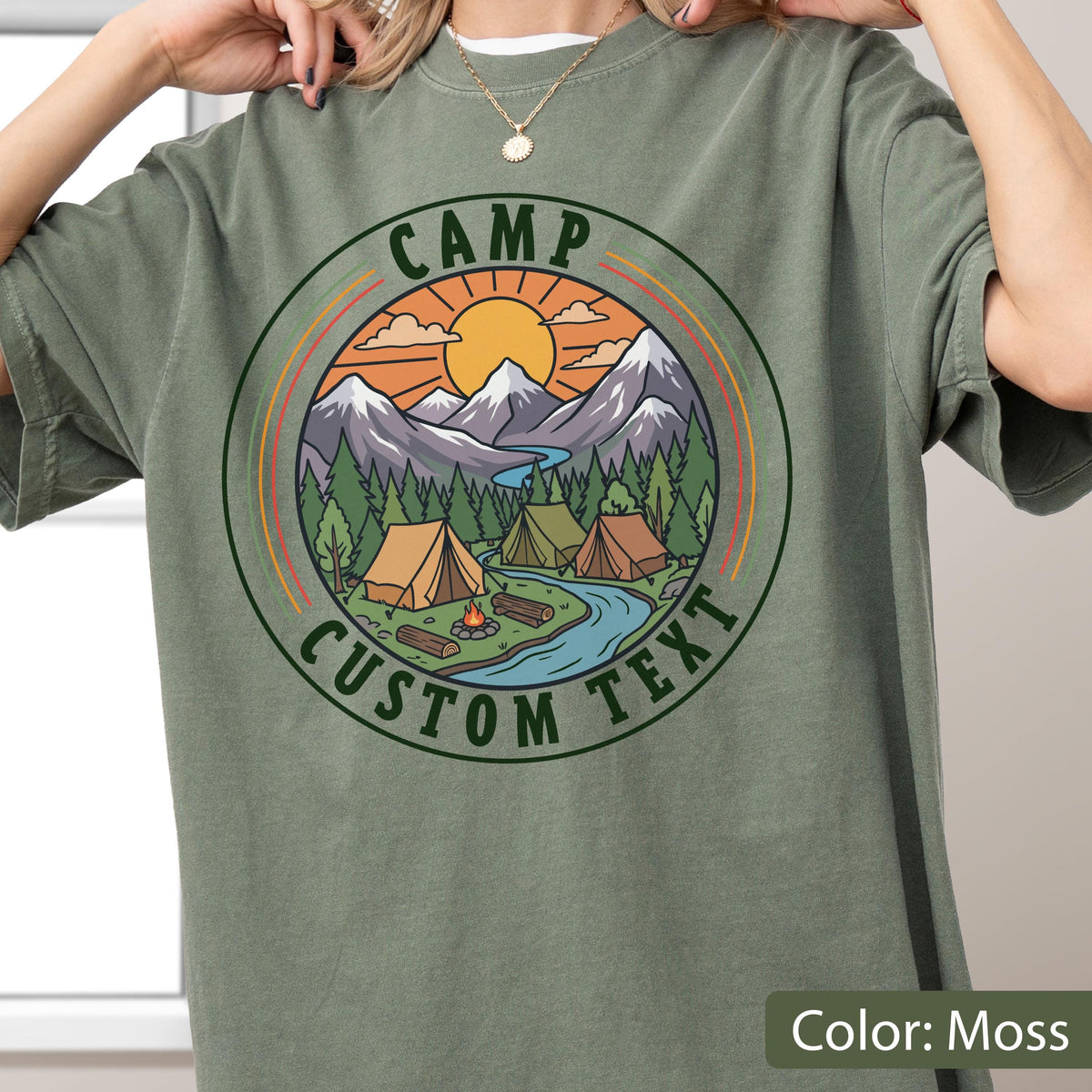 May include: A moss green t-shirt featuring a circular graphic with a mountain landscape, a sun, pine trees, tents, a campfire, and a river. The text "CAMP" is arched at the top and "CUSTOM TEXT" is arched at the bottom within the circular border.