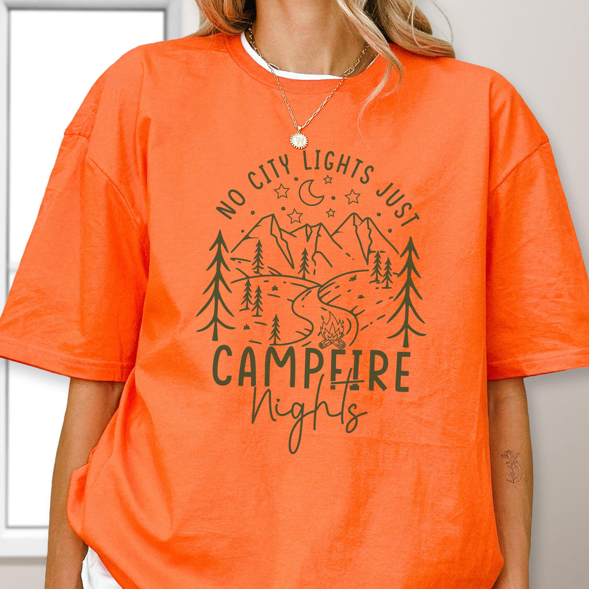 May include: An orange short-sleeve t-shirt featuring a dark green graphic print of a mountain landscape with pine trees, a crescent moon, stars, and a campfire. The text reads NO CITY LIGHTS JUST CAMPFIRE NIGHTS in a mix of block and script fonts.
