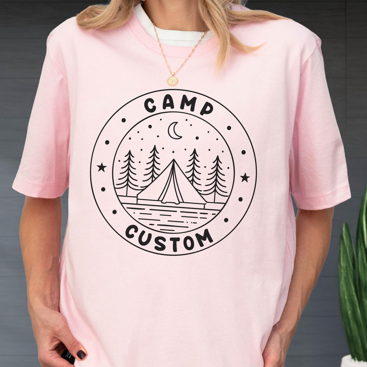 May include: A light pink short-sleeved t-shirt featuring a black circular graphic on the chest. The design includes a tent, pine trees, a crescent moon, and stars, with the text CAMP CUSTOM printed in a bold, rounded font within the circle border.