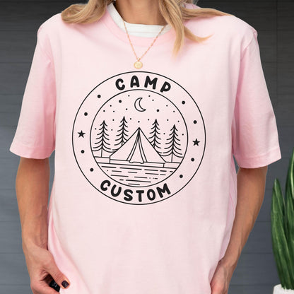 May include: A light pink short-sleeved t-shirt featuring a black circular graphic on the chest. The design includes a tent, pine trees, a crescent moon, and stars, with the text CAMP CUSTOM printed in a bold, rounded font within the circle border.
