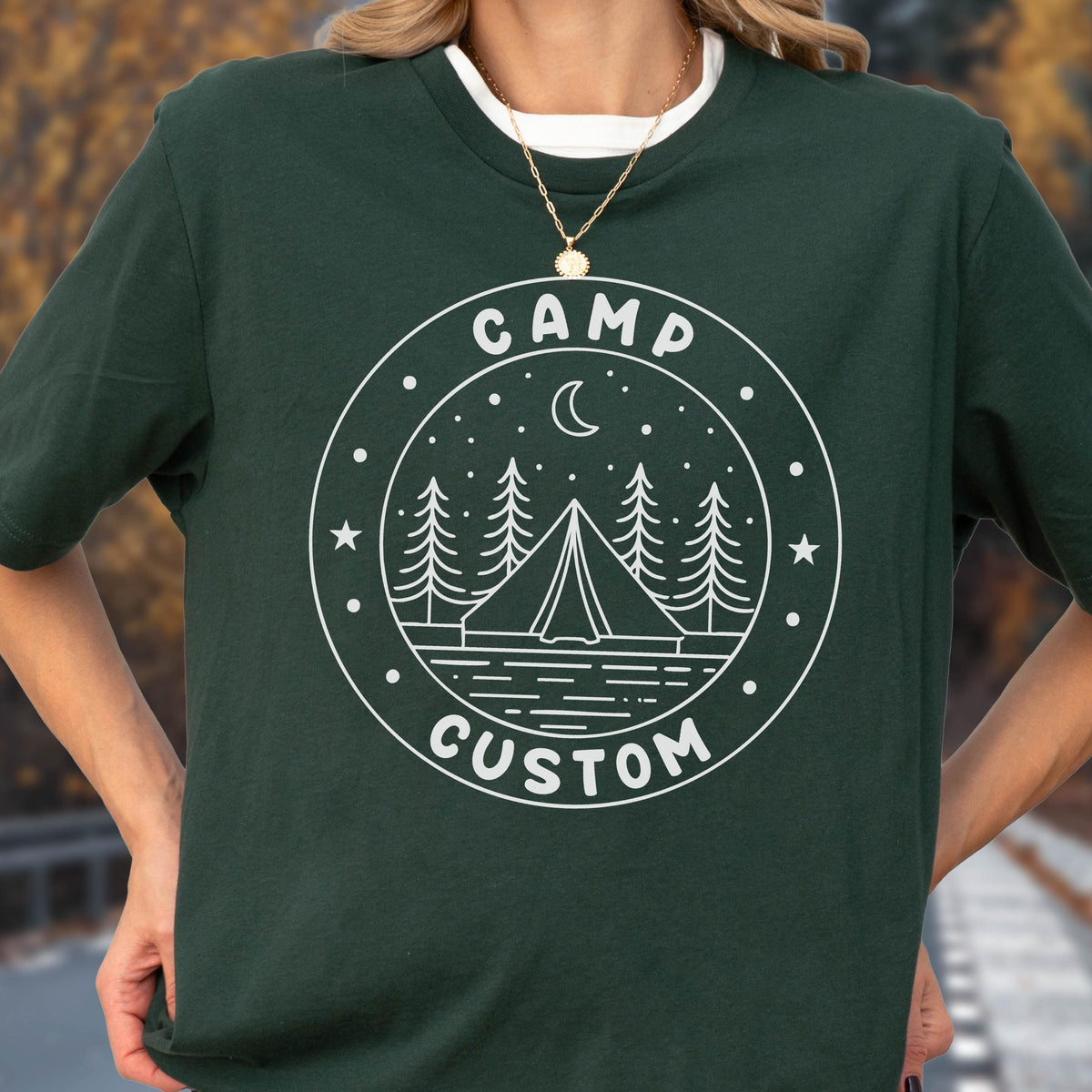 May include: A dark forest green short-sleeved t-shirt featuring a white circular graphic print. The design depicts a tent, pine trees, a crescent moon, and stars, with the text "CAMP" at the top and "CUSTOM" at the bottom of the circle.